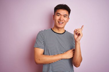 Asyalı genç bir Çinli, çizgili tişört giyiyor. İzole edilmiş pembe arka planda duruyor. Yüzünde kocaman bir gülümseme var..
