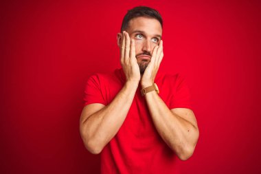 Kırmızı izole edilmiş arka plan üzerine gündelik tişört giyen genç yakışıklı. Yüzü, depresyonu ve üzüntüyü örten yorgun eller.