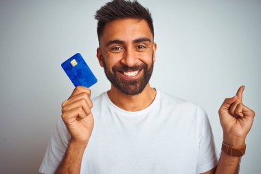 Young indian customer man holding credit card standing over isolated white background very happy pointing with hand and finger to the side