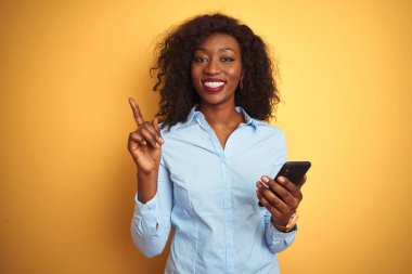 Genç Afrikalı Amerikalı kadın, izole edilmiş sarı arka planda duran akıllı telefonu kullanıyor. Bir fikir ya da soru işareti ile şaşırıyor.