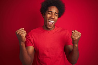 Afro saçlı genç Amerikalı adam izole edilmiş kırmızı arka planda dikilmiş mutlu ve heyecanlı bir şekilde kollarını kaldırıp, gülümseyerek ve başarı için çığlık atarak zafer hareketi yapıyor. Kutlama konsepti.