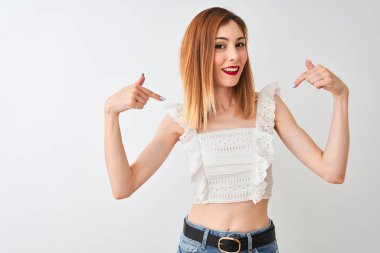 Izole beyaz arka plan üzerinde duran rahat t-shirt giyen güzel kızıl saçlı kadın yüzünde gülümseme ile emin görünümlü, parmakları gururlu ve mutlu kendini işaret.