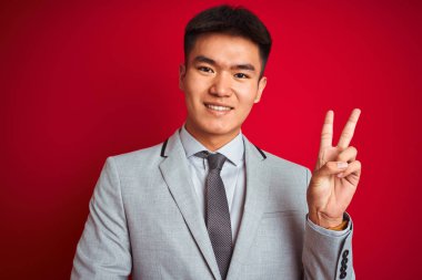 Gri ceketli ve kravatlı Asyalı Çinli işadamı izole edilmiş kırmızı arka planda dikiliyor ve kameraya gülümsüyor zafer işareti yapıyor. İki numara..