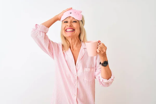 Middle age woman wearing sleep mask pajama drinking coffee over isolated white background stressed with hand on head, shocked with shame and surprise face, angry and frustrated. Fear and upset for mistake.