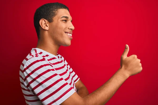 孤立した赤い背景にストライプのTシャツを着た若いハンサムなアラブ人男性誇りに思って 笑顔で親指を横にジェスチャー — ストック写真