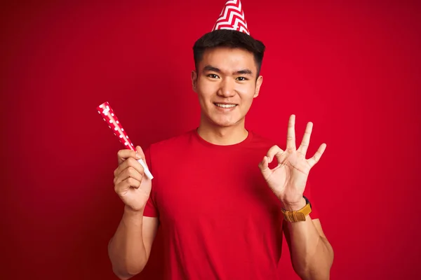 Asian chinese man on birthday celebration wearing funny hat over isolated red background doing ok sign with fingers, excellent symbol