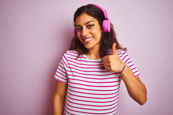 隔離されたピンクの背景の上のヘッドフォンを使用して音楽を聞く若い美しい女性は手で幸せな親指をジェスチャーする 成功を示すカメラを見て表現を承認する — ストック写真