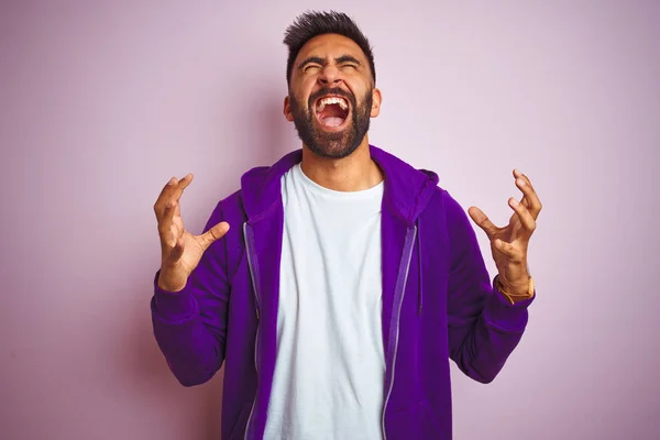 孤立したピンクの背景の上に立つ紫色のスウェットシャツを着た若いインド人男性は 狂気と怒りの叫びと積極的な表現と腕を上げて叫びました 欲求不満の概念 — ストック写真