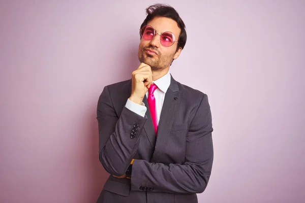 若いハンサムなビジネスマンは ピンクの背景の上にスーツとサングラスを着用し あごの質問 集中的な表現について考えて手で考えます 思慮深い顔で微笑んだ 疑いの概念 — ストック写真