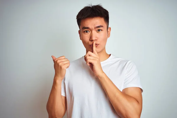 年轻的亚裔男子穿着T恤衫站在孤立的白色背景之上 要求保持安静 手指放在嘴唇上 手拉手指向侧面 沉默和秘密概念 — 图库照片