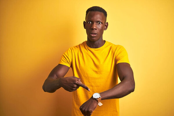 Young african american man wearing casual t-shirt standing over isolated yellow background In hurry pointing to watch time, impatience, upset and angry for deadline delay