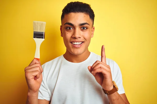 Young brazilian painter man holding brush standing over isolated yellow background surprised with an idea or question pointing finger with happy face, number one