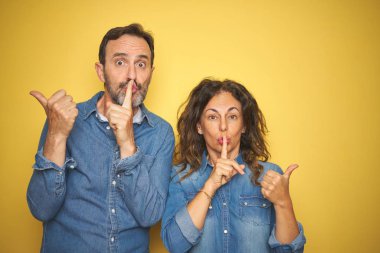 Beautiful middle age couple together standing over isolated yellow background asking to be quiet with finger on lips pointing with hand to the side. Silence and secret concept. clipart