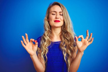Mavi izole edilmiş arka planda duran genç ve güzel bir kadın rahatla ve gözün kapalı gülümse parmaklarınla meditasyon hareketi yap. Yoga konsepti.