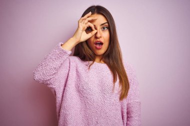 İzole edilmiş pembe arka plan üzerinde duran sıradan süveter giyen genç ve güzel bir kadın şaşırmış bir yüz ve parmaklarından bakan bir gözle şaşırmış bir jest yapıyor. İnançsız ifade.