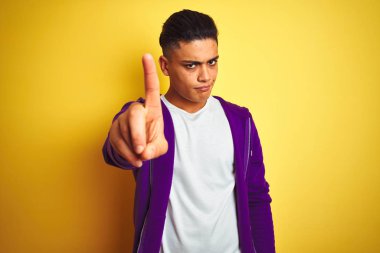 Mor kazak giyen genç Brezilyalı adam izole edilmiş sarı arka planda duruyor. Parmağını kaldırıyor ve kızgın bir ifade sergiliyor.