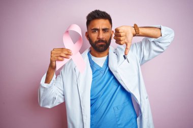 Young indian doctor man holding cancer ribbon standing over isolated pink background with angry face, negative sign showing dislike with thumbs down, rejection concept