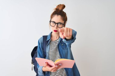 Redhead student woman wearing backpack reading book over isolated white background pointing with finger to the camera and to you, hand sign, positive and confident gesture from the front