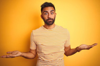 İzole edilmiş sarı arka planda duran tişörtlü genç Hintli adam hiçbir şeyden habersiz ve kolları açık, hiçbir fikri yok..