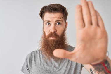 Genç, kızıl saçlı, tişörtlü, izole edilmiş gri arka planda duran, ciddi ve kendinden emin ifadesiyle dur işareti yapan, savunma jesti