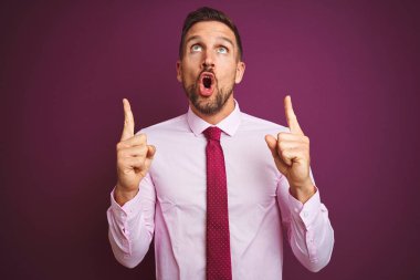Zarif gömlek ve kravat giyen genç iş adamı, mor izole arka planda şaşkın ve şaşırmış bir şekilde yukarı bakıyor ve parmaklarıyla işaret ediyor ve kollarını kaldırıyordu..