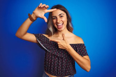Young beautiful woman wearing floral t-shirt over blue isolated background smiling making frame with hands and fingers with happy face. Creativity and photography concept. clipart