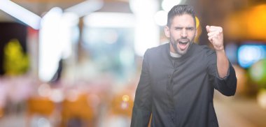 Young Christian priest over isolated background angry and mad raising fist frustrated and furious while shouting with anger. Rage and aggressive concept.