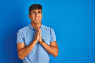 Sıradan polo giyen genç Hintli adam izole edilmiş mavi arka planda duruyor. Elleriyle yalvarıyor ve dua ediyor. Yüzünde umut ifadesi var. Af diliyor. Din kavramı.
