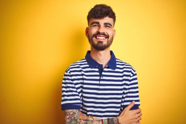 İzole edilmiş sarı arka planda duran çizgili polo giyen genç adam kollarını kavuşturup kameraya bakarken gülümsüyor. Pozitif kişi.