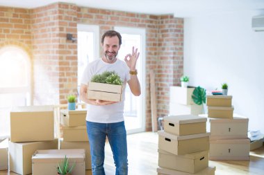Orta yaşlı bir adam yeni bir eve taşınıyor. Karton kutular paketliyor. Tamam işareti yapıyor.