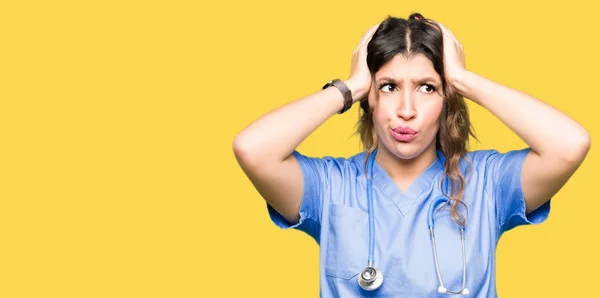 医療ユニフォームクレイジーを着て 頭の上に手で怖がって 口を開いてショックを恐れ 驚いた若い成人医師の女性 — ストック写真