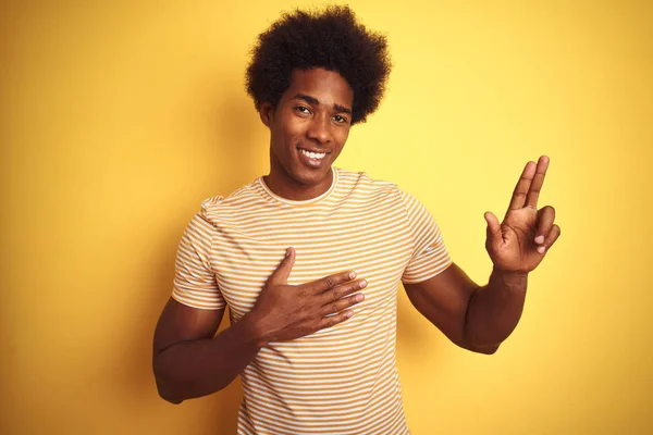 American man with afro hair wearing striped t-shirt standing over isolated yellow background smiling swearing with hand on chest and fingers up, making a loyalty promise oath