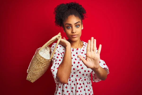 African american woman wearing fashion dress holding wicker bag over isolated red background with open hand doing stop sign with serious and confident expression, defense gesture