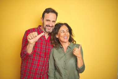 Izole sarı arka plan üzerinde güzel orta yaş çift mutlu yüz görünümlü ve başparmak ile yan işaret gülümseyerek.