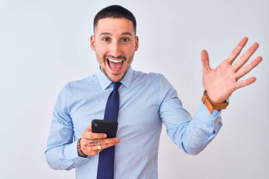 Genç yakışıklı iş adamı izole edilmiş arka planda akıllı telefon kullanıyor çok mutlu ve heyecanlı, kazanan yüz ifadesi zaferi büyük bir gülümsemeyle kutluyor ve ellerini kaldırıyor.
