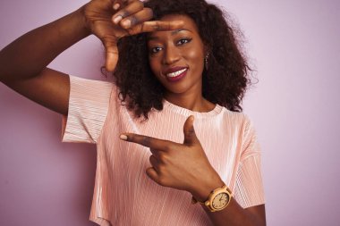 Genç Afro-Amerikan kadın tişört giyiyor pembe arka planda duruyor gülümsüyor ve elleri ve parmakları mutlu yüzlü bir çerçeve yapıyor. Yaratıcılık ve fotoğrafçılık kavramı.