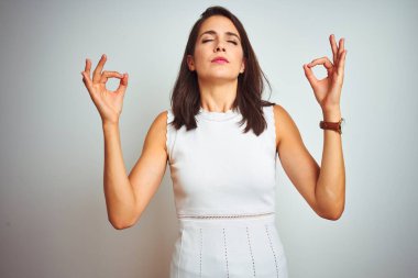 İzole edilmiş beyaz arka plan üzerinde duran elbiseler giyen genç ve güzel bir kadın rahatlamış ve parmaklarıyla meditasyon hareketi yaparken gözleri kapalı gülümsüyor. Yoga konsepti.