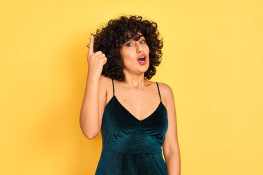 Sarı arka planda zarif elbiseler giyen kıvırcık saçlı genç Arap kadın başarılı bir fikri işaret ediyor. Heyecanlı ve mutlu. Bir numara..