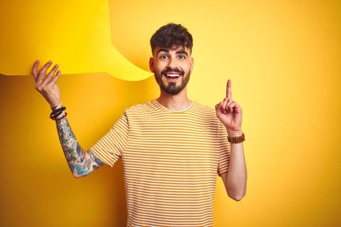 Dövmeli genç adam konuşma baloncuğu tutarak izole edilmiş sarı arka planda duruyor. Bir fikir ya da soru işareti ile şaşırmış, mutlu bir yüzle, bir numara.