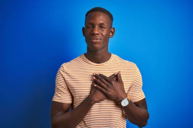 Siyah çizgili günlük tişört giyen Afro-Amerikan bir adam izole edilmiş mavi arka plan üzerinde duruyor elleri göğsünde kapalı ve yüzünde minnet dolu bir jest ile gülümsüyor. Sağlık kavramı.