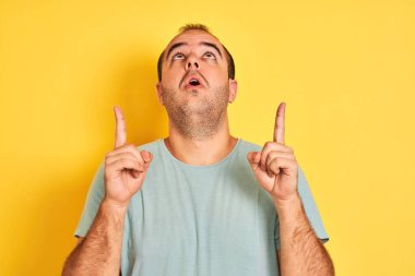 Yeşil tişörtlü genç adam, izole edilmiş sarı arka planda duruyor. Şaşırmış ve şaşırmış bir şekilde yukarı bakıyor ve parmaklarıyla işaret ediyor ve kollarını kaldırıyor..