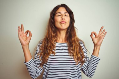 İzole edilmiş beyaz arka plan üzerine çizgili tişört giyen genç ve güzel bir kadın rahatla ve parmaklarıyla meditasyon hareketi yaparken gözleri kapalı gülümse. Yoga konsepti.
