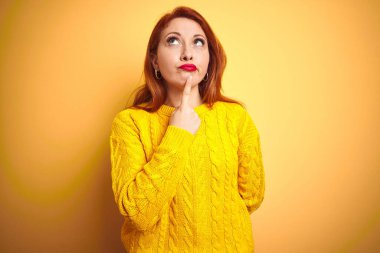 Kışlık süveter giyen güzel kızıl saçlı kadın izole edilmiş sarı arka planda dikilirken parmakları çenesinde olan şüpheyi düşünüp kafasını kaldırıyor.