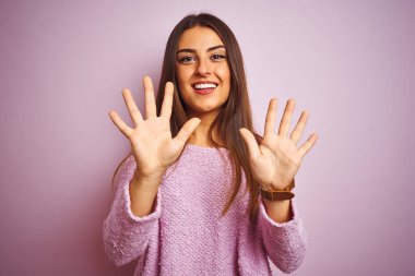 İzole edilmiş pembe arka plan üzerinde duran sıradan bir süveter giyen genç ve güzel bir kadın on numarayı gösterirken kendinden emin ve mutlu bir şekilde gülümsüyor..