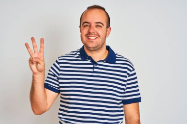 Sıradan çizgili polo giyen genç adam izole edilmiş beyaz arka planda duruyor ve üç numaralı parmaklarıyla işaret ederken kendinden emin ve mutlu bir şekilde gülümsüyor..