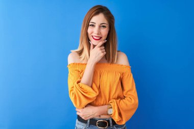 Güzel kızıl saçlı kadın turuncu günlük tişört giyiyor mavi arka planda dikiliyor kameraya bakıyor kollarını kavuşturmuş ve çenesini kaldırmış gülümsüyor. Olumlu düşünüyorum..