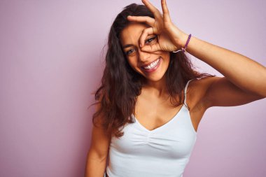 İzole edilmiş pembe arka plan üzerinde duran beyaz tişört giyen genç ve güzel bir kadın el hareketleriyle gülümsüyor ve mutlu yüzle parmaklarının arasından bakıyor..