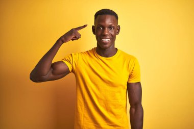 Genç Afro-Amerikalı bir adam günlük tişört giyip izole edilmiş sarı arka planda dikiliyor. Tek parmakla kafasını göstererek gülümsüyor. Harika bir fikir ya da düşünce, iyi bir hafıza.
