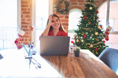 Güzel bir kadın evde dizüstü bilgisayarla oturuyor Noel ağacının etrafında el hareketleriyle gülümsüyor ve mutlu yüzüyle parmaklarının arasından bakıyor..