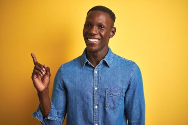 Kotlu gömlek giyen genç Afro-Amerikalı adam izole edilmiş sarı arka planda duruyor. Yüzünde kocaman bir gülümseme, eli ve parmağı kameraya bakıyor..